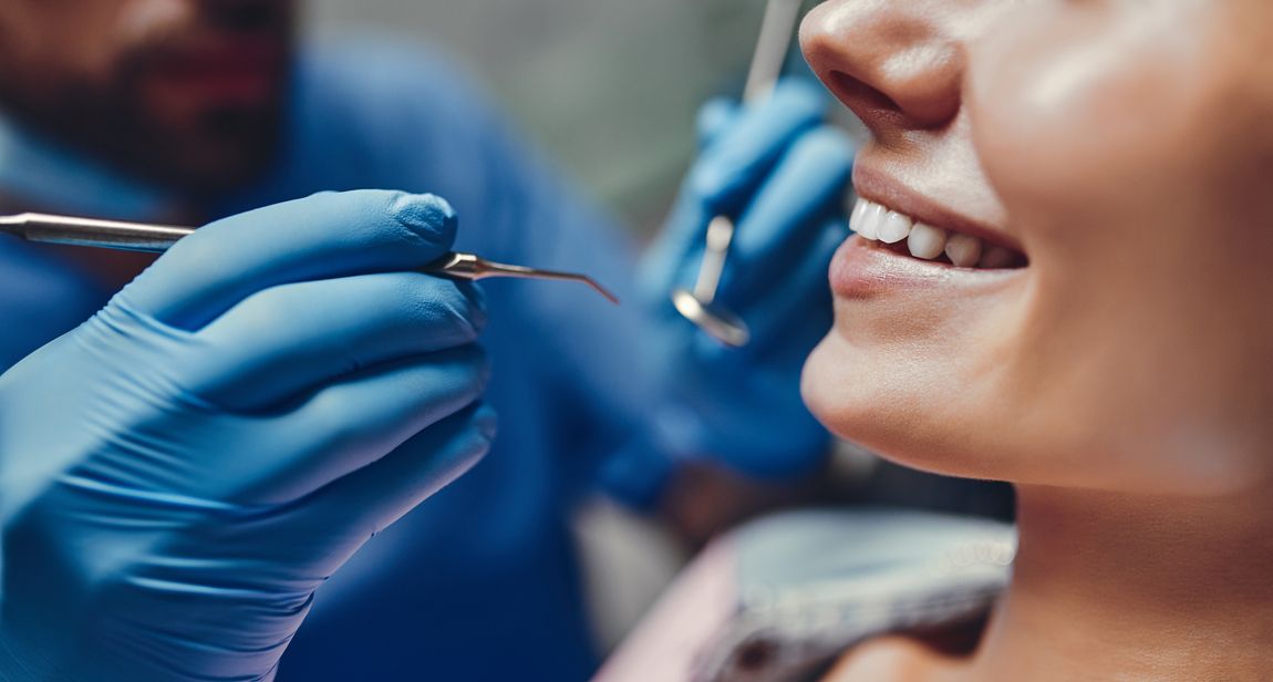 Ein Zahnarzt in blauen Handschuhen und Uniform untersucht mit dentalen Instrumenten das Lächeln einer Patientin, Nahaufnahme des Mundbereichs