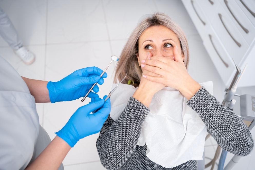 Eine Patientin mit ängstlichem Gesichtsausdruck, die den Mund mit den Händen bedeckt, während ein Zahnarzt in blauen Handschuhen und Uniform mit dentalen Instrumenten arbeitet, im Hintergrund eine zahnmedizinische Praxis